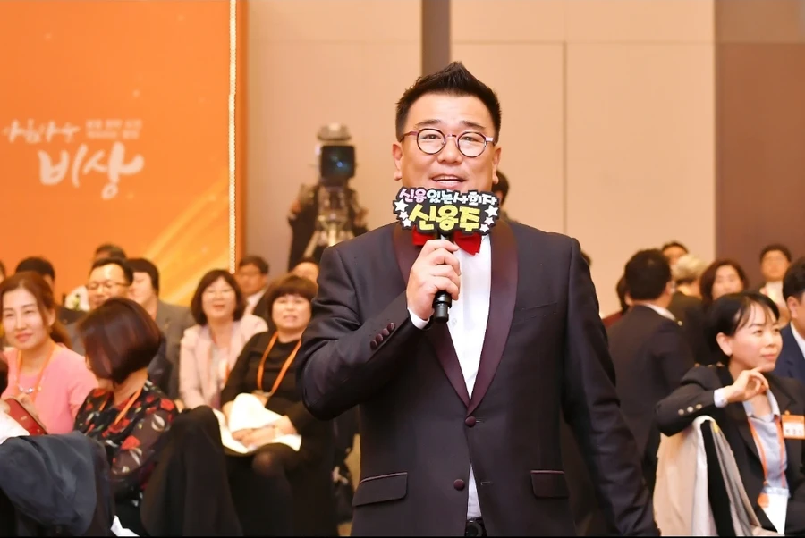 정장에 빨간 나비넥타이를 매고 이름팻말을 든 채 청중 앞에서 마이크를 들고 있는 사회자