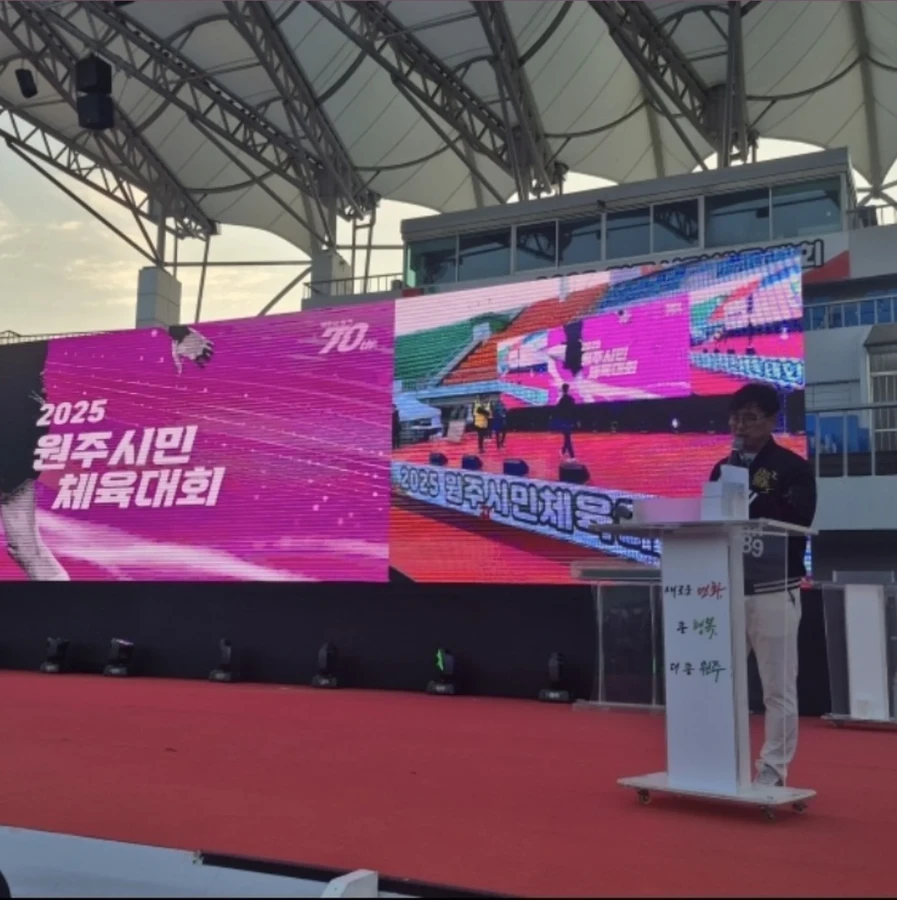 레드카펫이 깔린 야외 무대 연단 옆에서 마이크로 연설하는 정장 차림 사회자