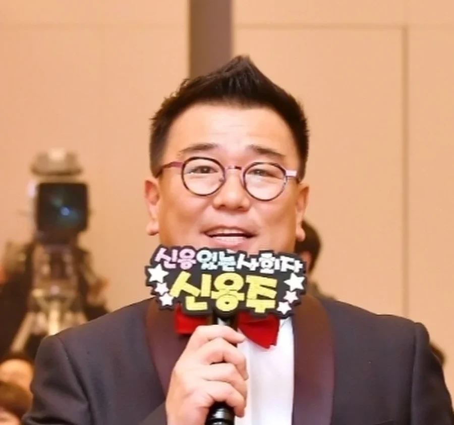 무대에서 나비넥타이를 맨 정장 차림으로 이름팻말과 마이크를 들고 있는 사회자 클로즈업