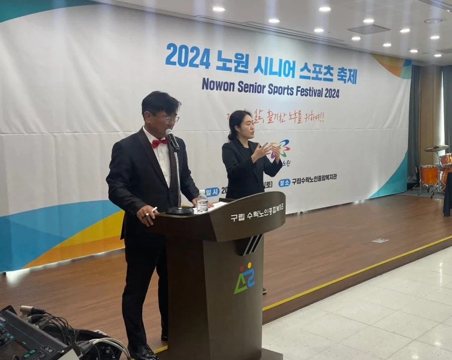 연단 뒤에서 정장을 입고 마이크로 진행하는 사회자와 통역하는 진행자가 있는 실내 행사 무대