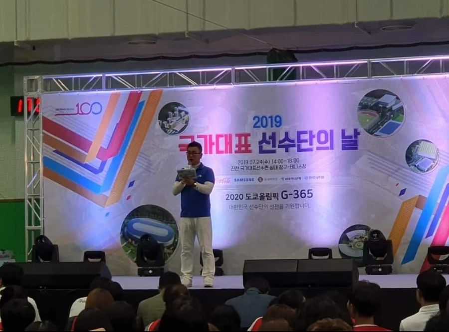 대형 배너 무대 위에서 서류를 들고 마이크로 진행하는 정장 차림 사회자