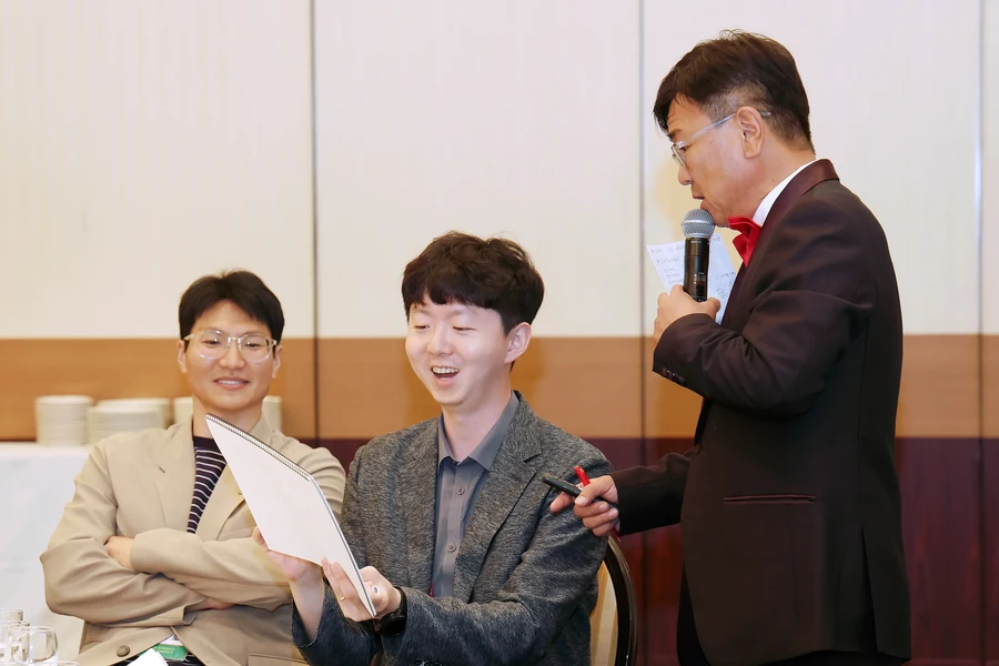 연회장 무대에서 마이크와 진행 자료를 든 정장 차림 사회자와 테이블에 둘러앉은 참석자들